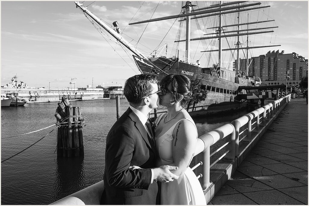 A bride and groom kiss on a sidewalk with Moshulu Philadelphia behind them.
