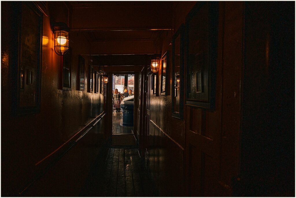 At the end of a dark hall, wedding guests gather in a dining room.
